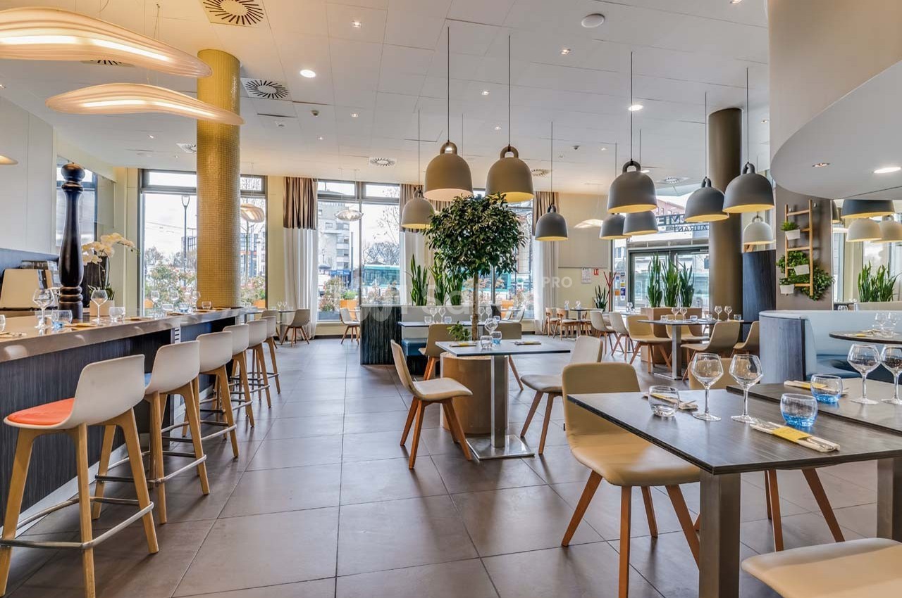 Salle de restaurant moderne avec tables dressées, chaises colorées, suspensions lumineuses et grandes baies vitrées.
