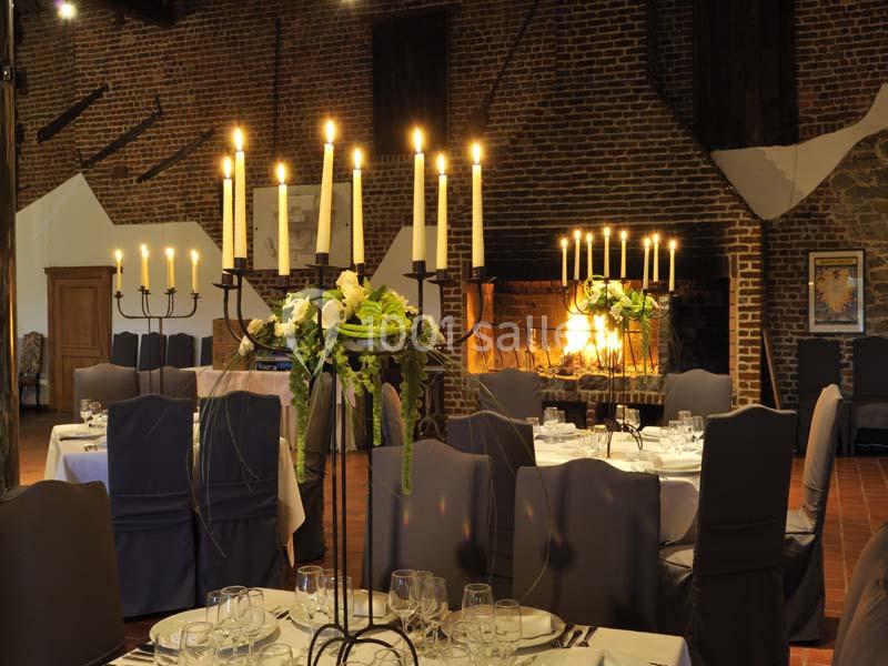 Salle de réception avec tables dressées, chandeliers allumés et cheminée en pierre au fond.