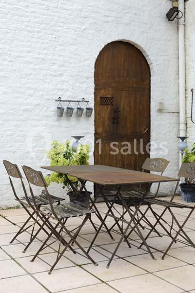 Table et chaises en métal sur une terrasse en dalles, devant un mur blanc avec une porte en bois et des plantes décoratives.
