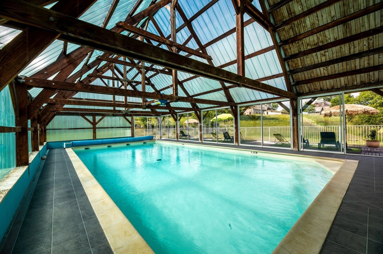 Piscine intérieure lumineuse sous une structure en bois avec toit en verre, entourée de chaises longues.