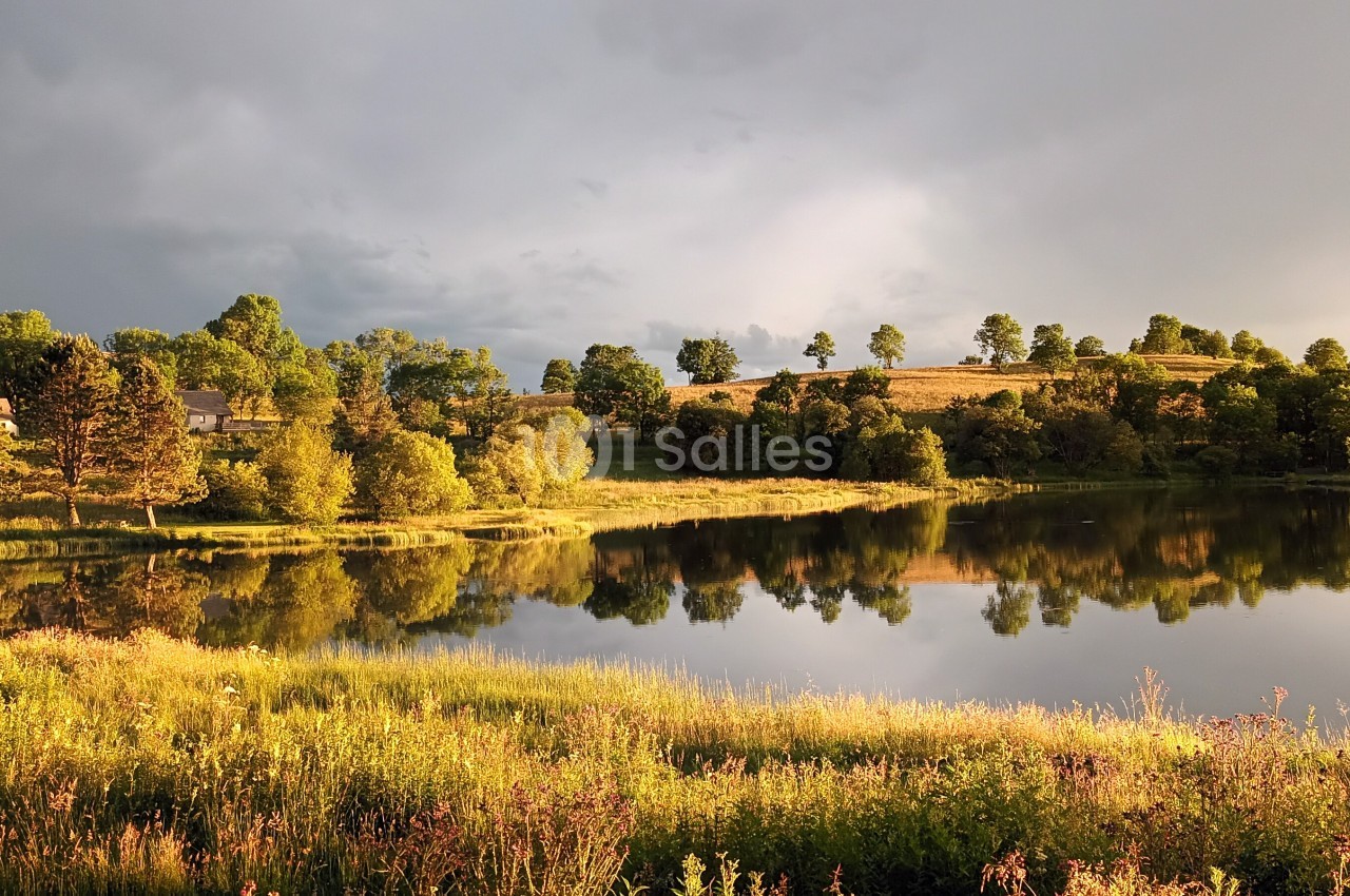 Paysage naturel avec un lac reflétant des arbres et des collines sous une lumière douce en fin de journée.