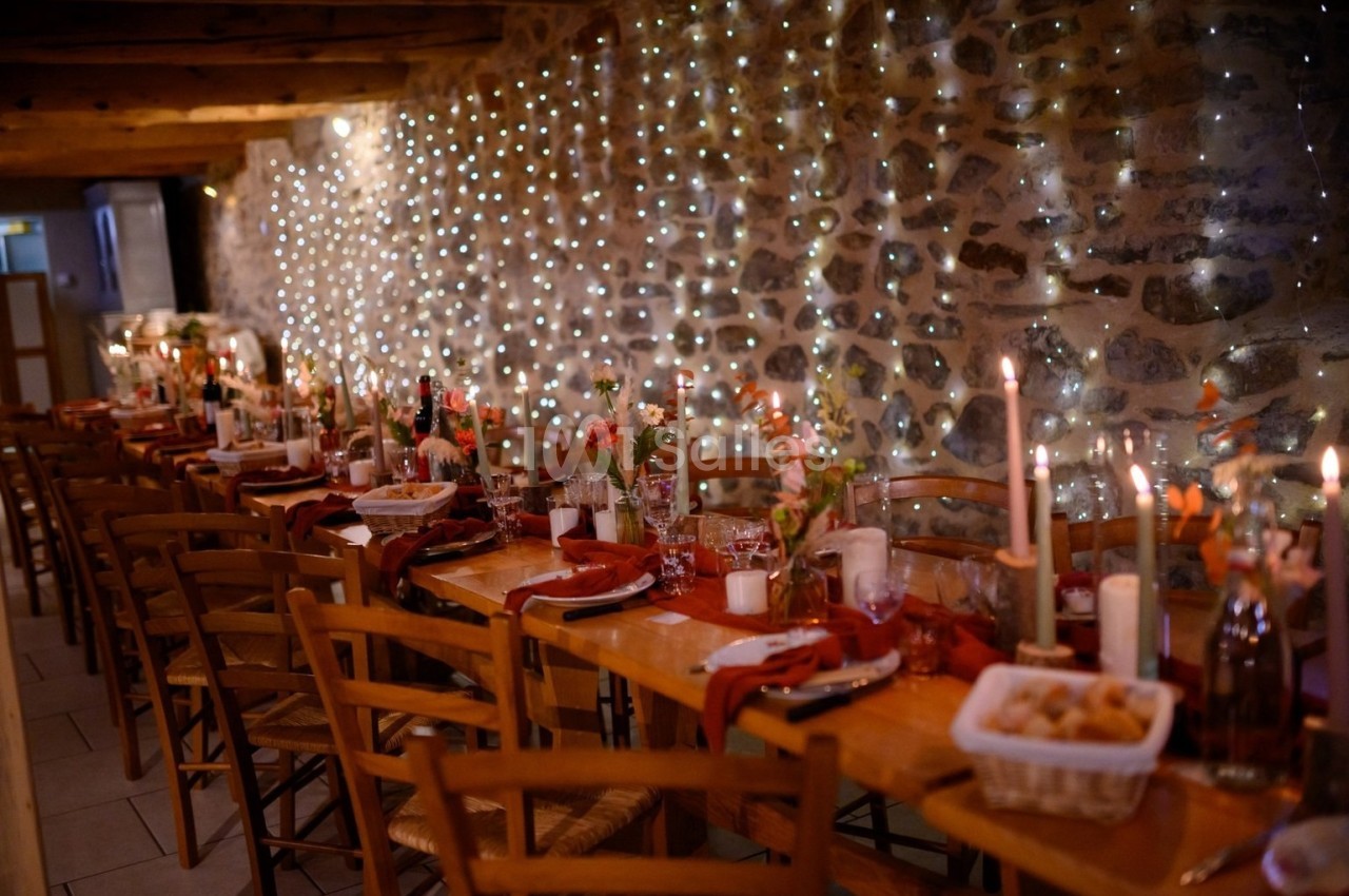 Salle rustique décorée pour un dîner, avec longues tables en bois, bougies allumées et guirlandes lumineuses sur un mur en…