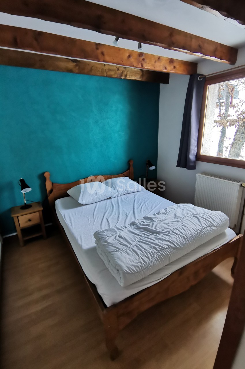 Chambre avec lit double en bois, mur turquoise, poutres apparentes, fenêtre donnant sur un arbre, parquet au sol.