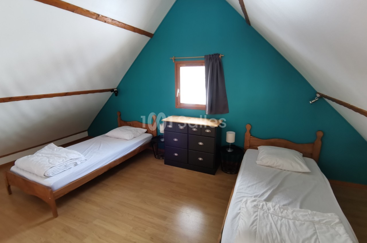 Deux lits simples avec draps blancs dans une chambre mansardée, murs turquoise et parquet clair.