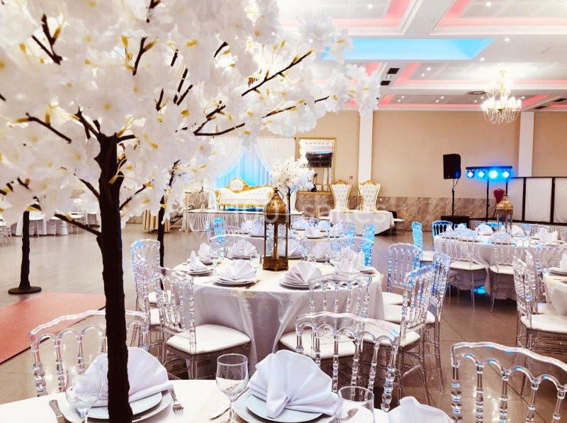 Salle de réception décorée avec des tables rondes, nappes blanches, chaises transparentes et arbres artificiels fleuris.