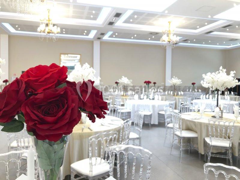 Salle de réception élégante avec tables dressées, chaises transparentes et décoration florale rouge et blanche.