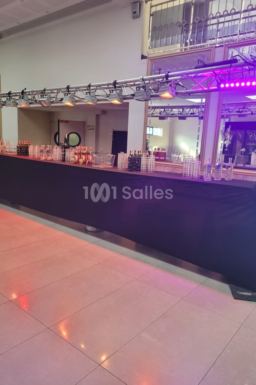 Bar long avec nappes noires, verres, bouteilles et éclairage violet dans une salle lumineuse avec miroirs.