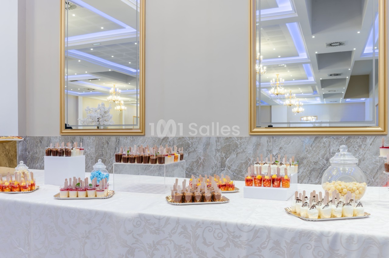 Table garnie de desserts variés en verrines et bonbons, disposée dans une salle élégante avec miroirs et lustres.