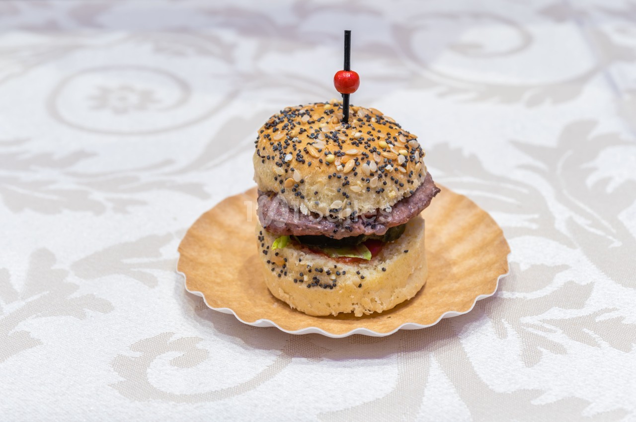 Mini burger sur un pain aux graines, garni de viande et de légumes, présenté sur une assiette en papier.