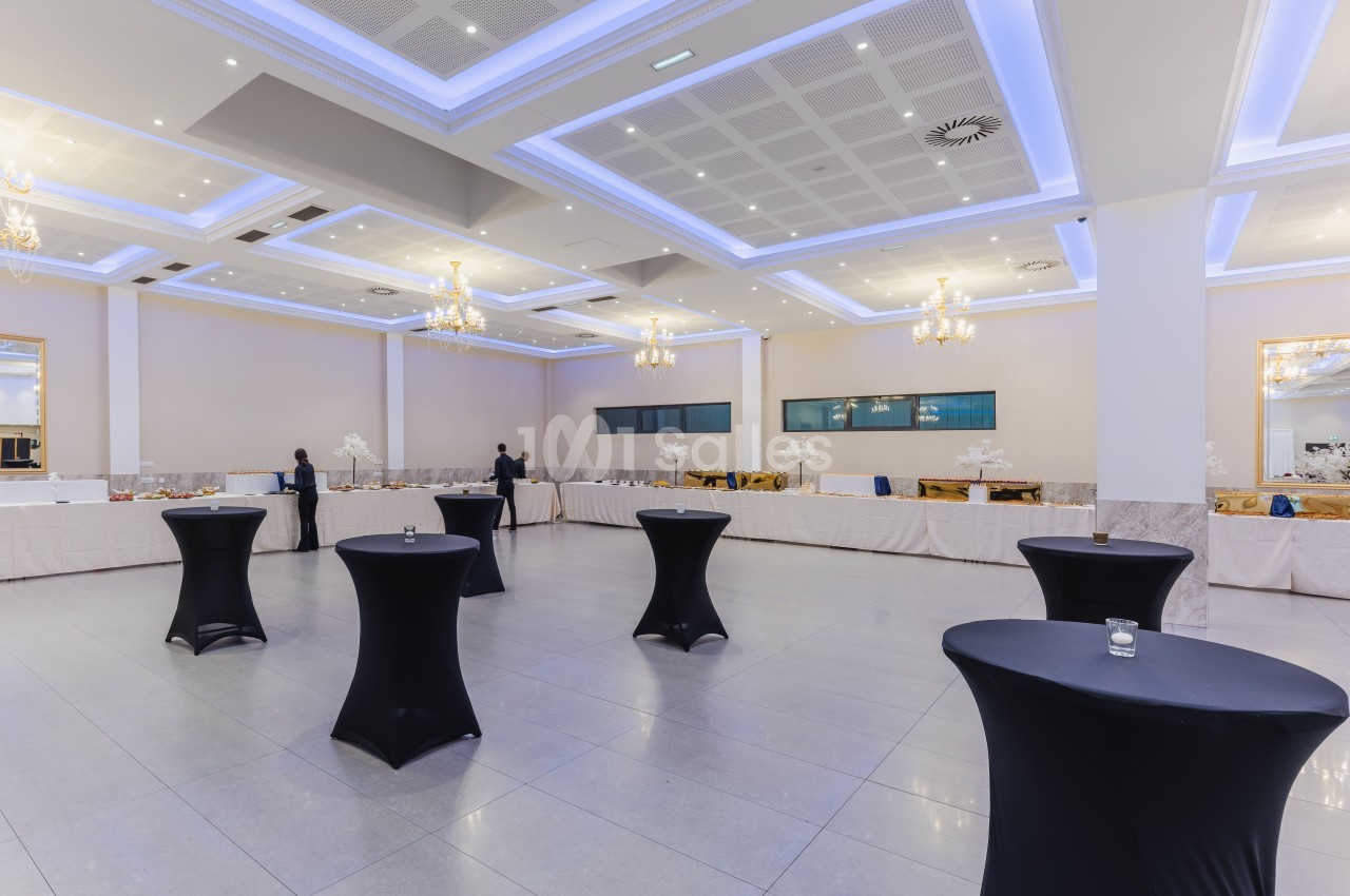 Salle de réception spacieuse avec tables hautes noires et buffet dressé le long des murs, éclairée par des lustres.