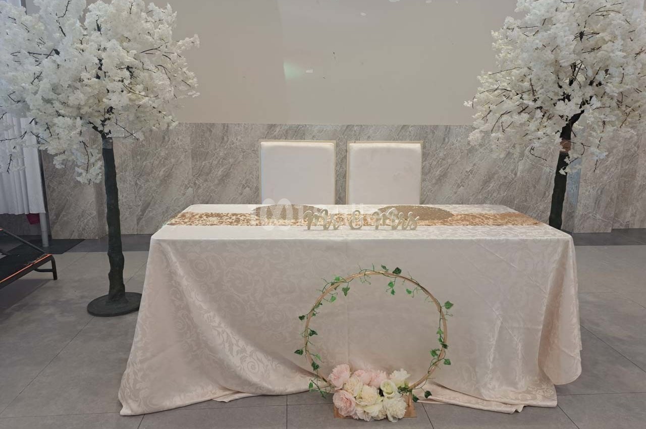 Table décorée pour un mariage avec nappe blanche, panneau ’Mr & Mrs’, fleurs et arbres artificiels en arrière-plan.