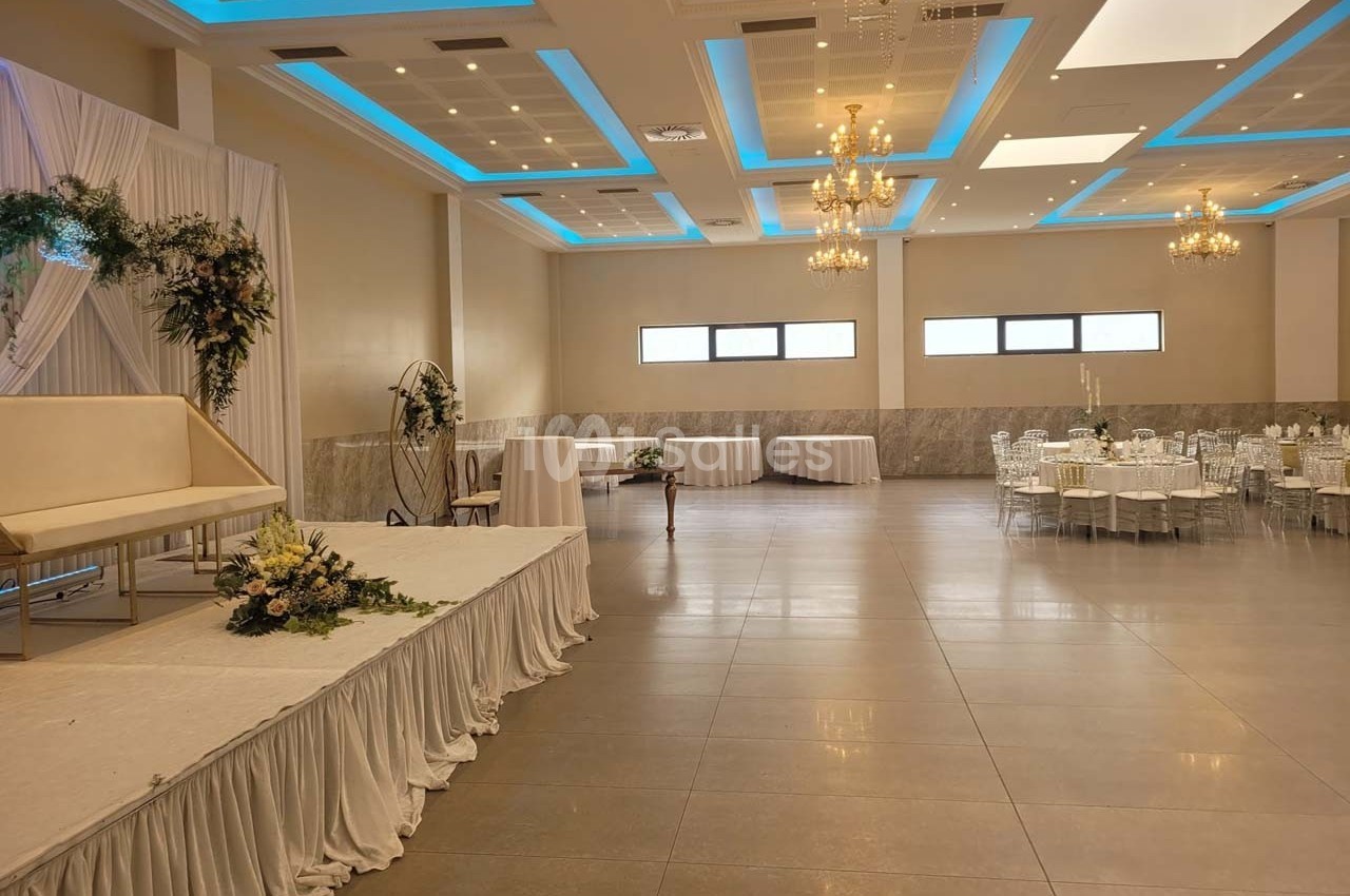 Salle de réception lumineuse avec tables dressées, scène décorée de fleurs et lustres suspendus au plafond.