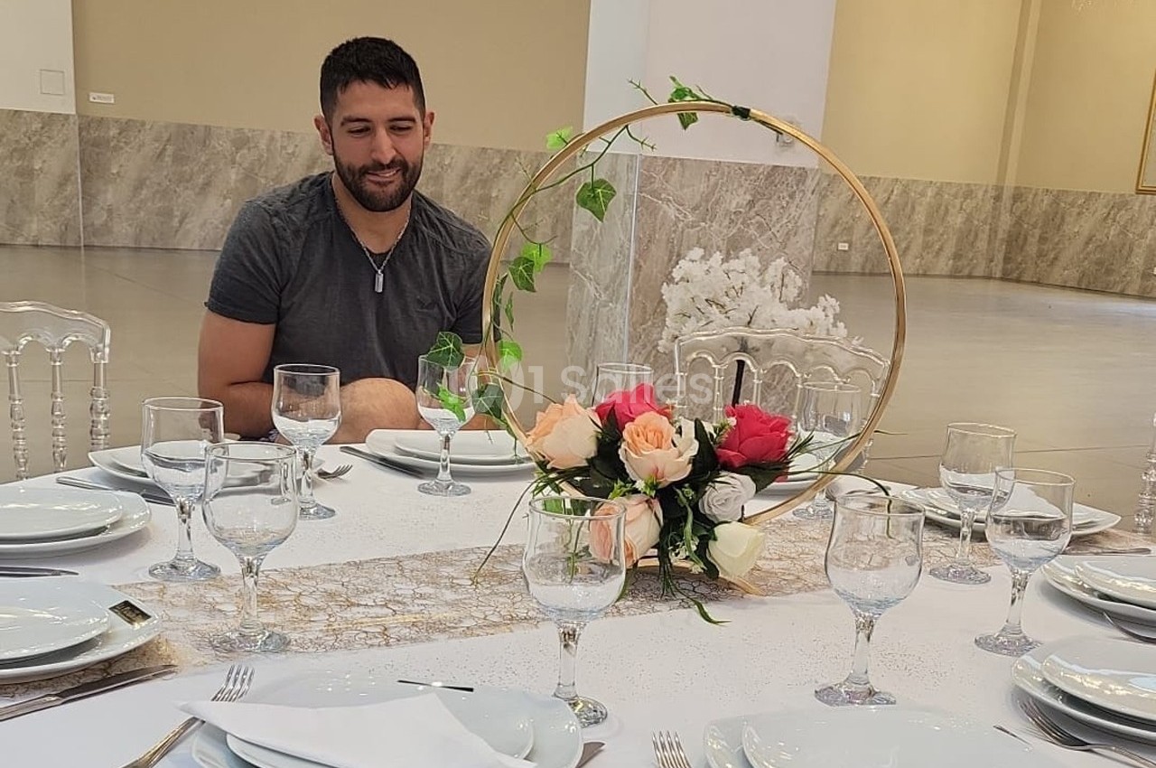 Un homme assis à une table élégamment dressée avec des fleurs et des verres, dans une grande salle lumineuse.