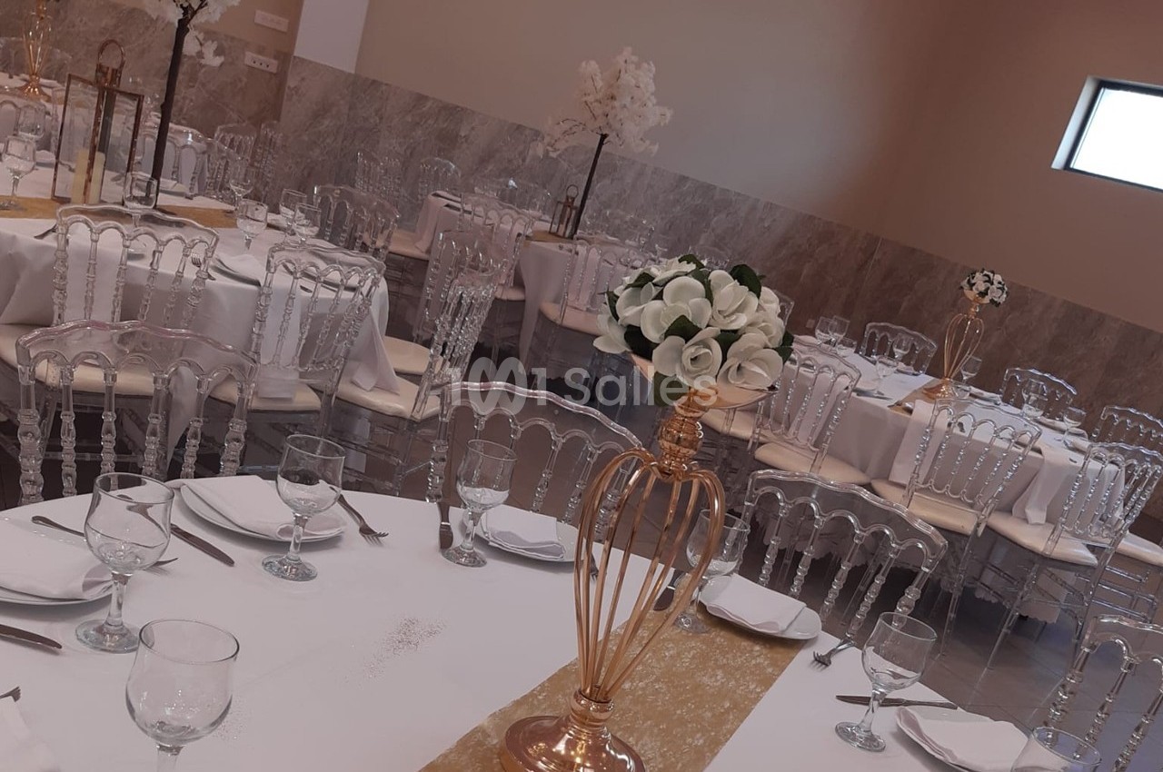 Salle de réception élégamment décorée avec des tables dressées, des chaises transparentes et des centres de table fleuris.