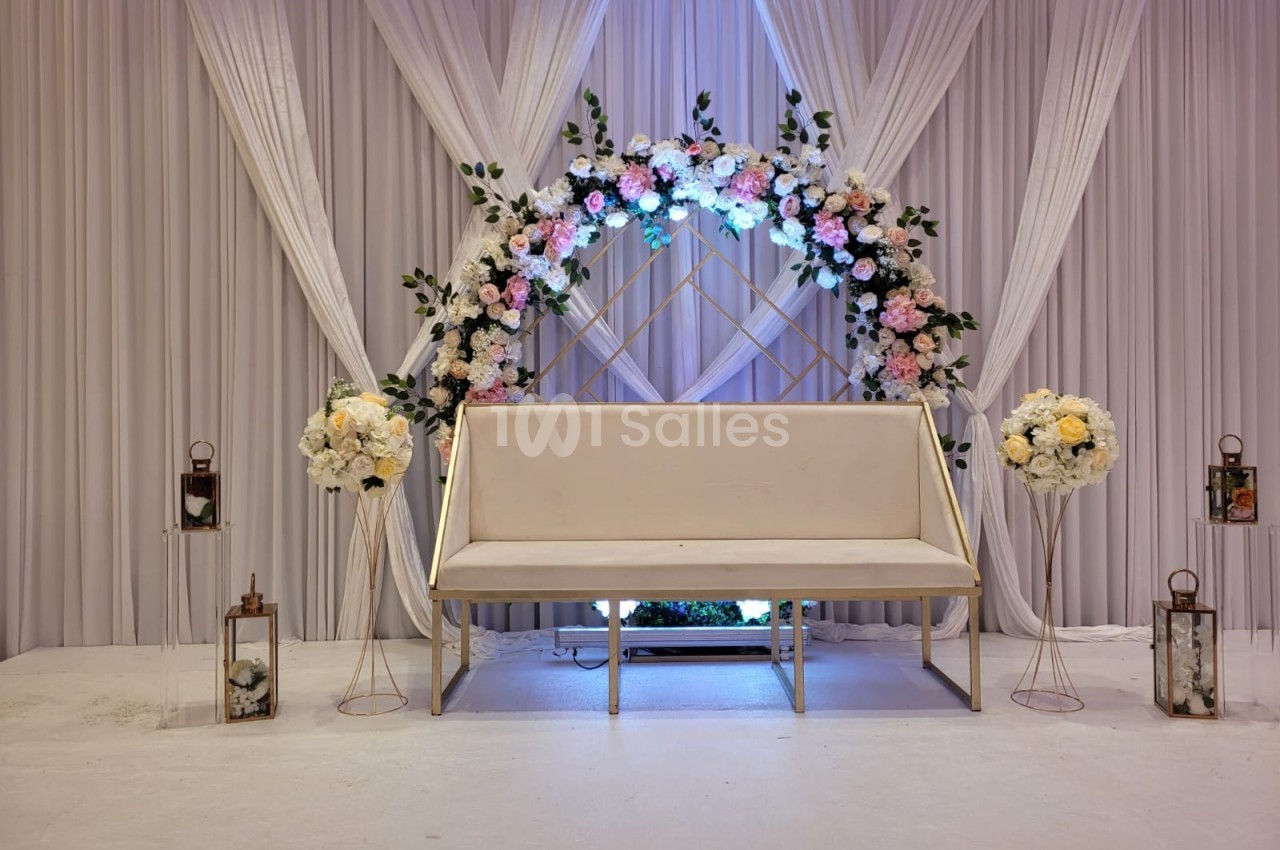 Banc beige devant une arche florale ornée de roses blanches et roses, avec des rideaux blancs en arrière-plan.