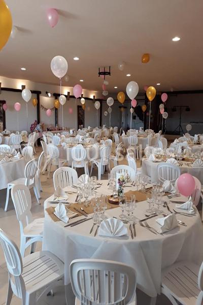 Salle décorée pour un événement avec des tables rondes dressées, des chaises blanches et des ballons colorés suspendus.