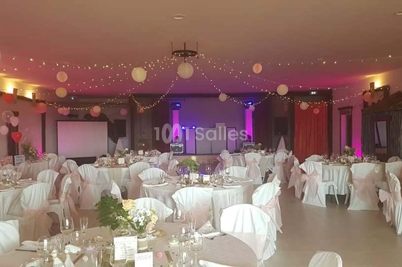 Salle de réception décorée avec des tables dressées, des chaises couvertes de housses blanches et des guirlandes lumineuses…