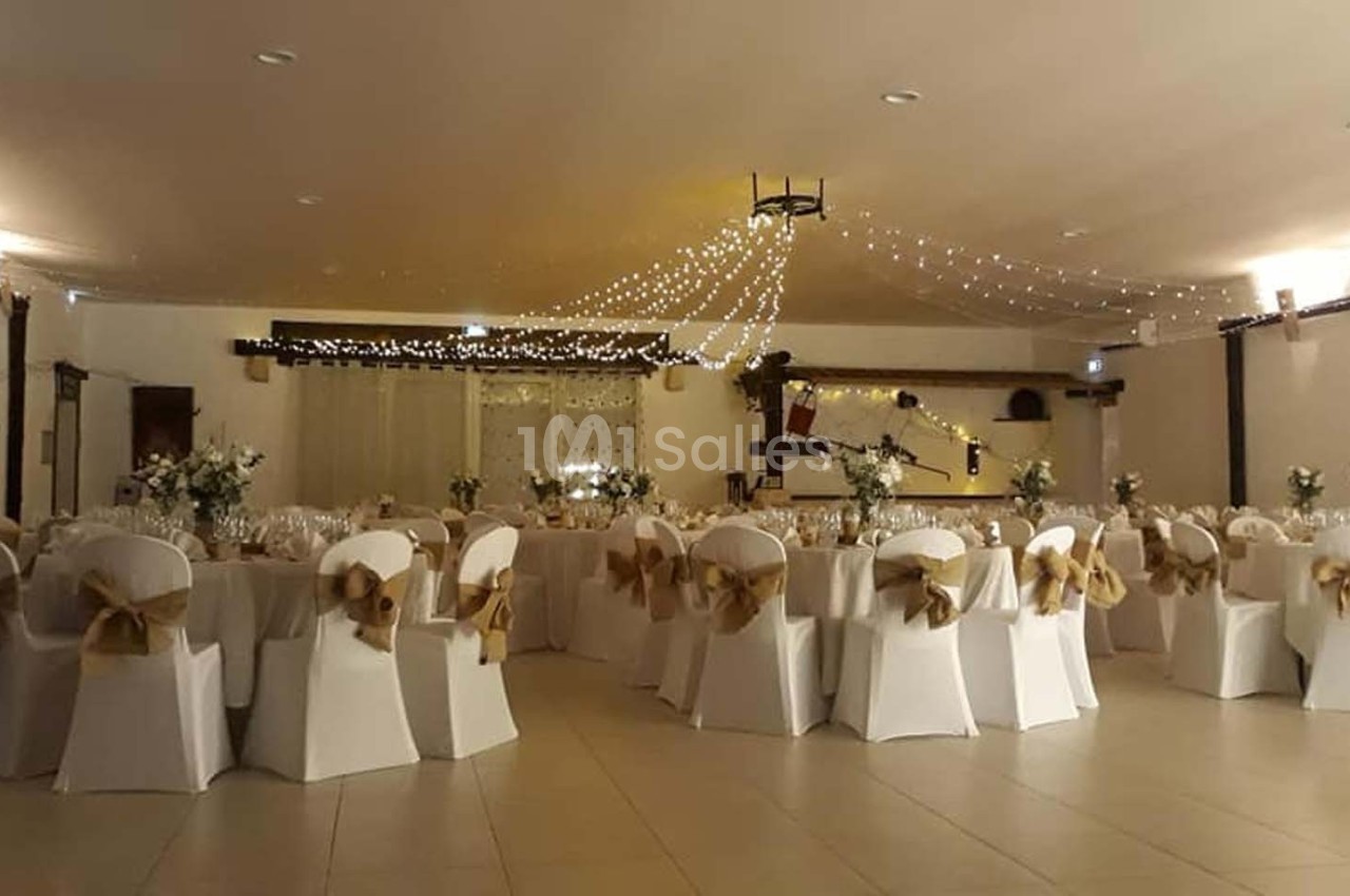 Salle de réception décorée avec des chaises blanches ornées de nœuds dorés et des guirlandes lumineuses suspendues.