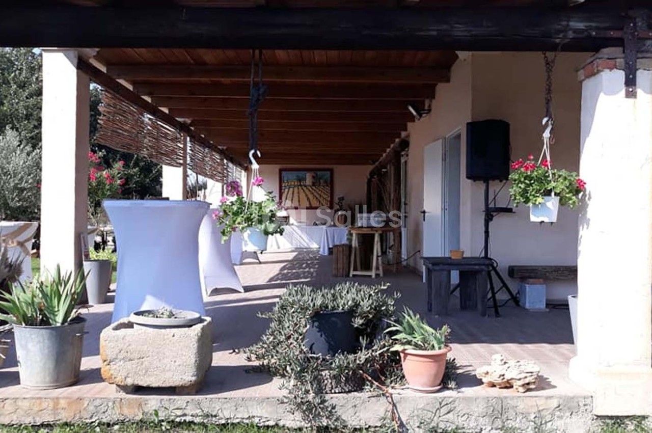 Terrasse couverte avec plantes en pots, table haute blanche et décoration suspendue, dans un cadre extérieur ensoleillé.