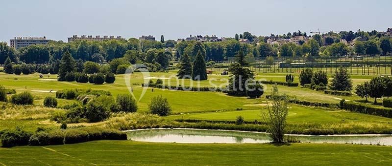 Vue d'un vaste terrain de golf verdoyant avec des arbres, un étang et des bâtiments en arrière-plan.