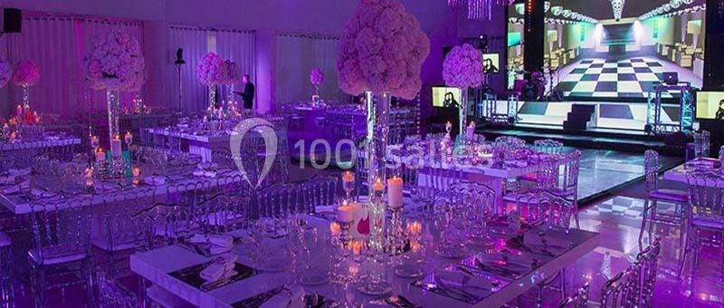 Salle de réception décorée avec des tables élégantes, des chandeliers et un éclairage coloré, avec une scène en arrière-plan.