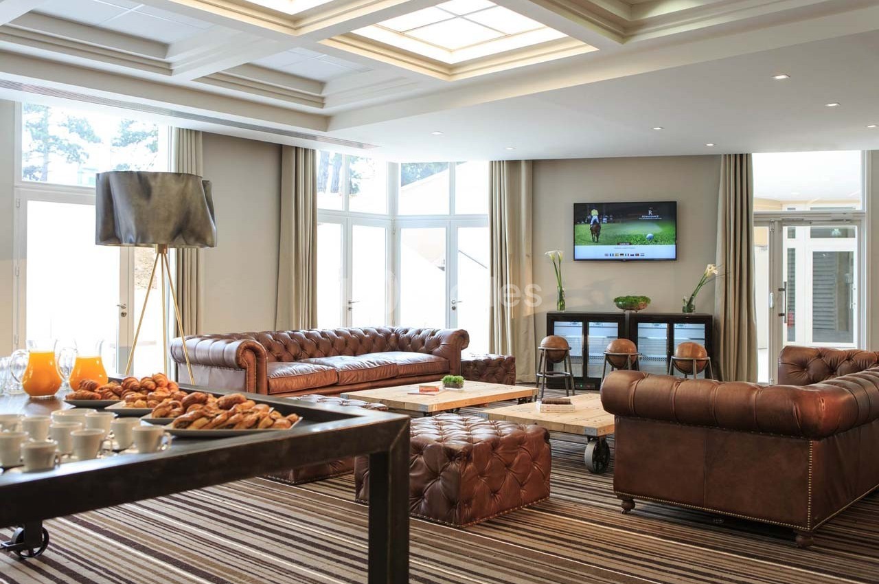 Salon lumineux avec canapés en cuir, table basse, écran mural allumé, et buffet avec boissons et viennoiseries.