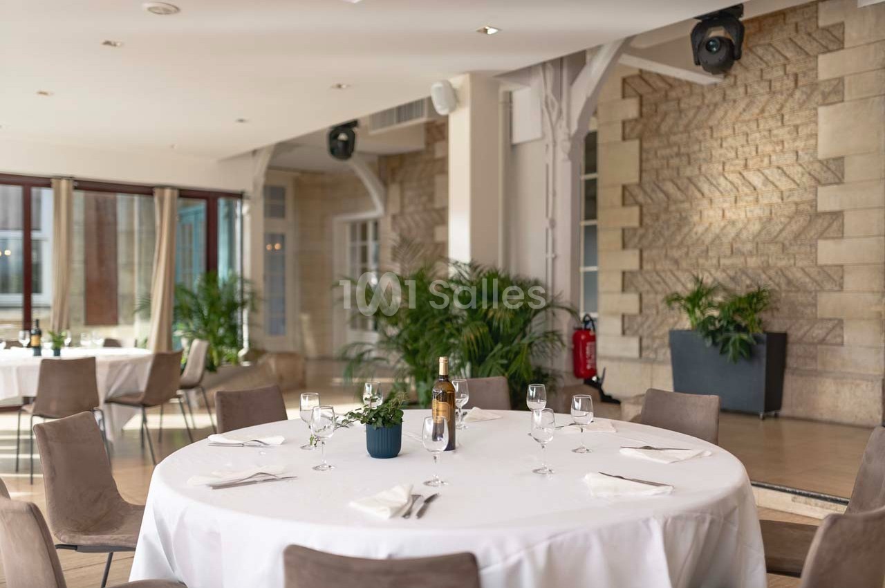 Salle lumineuse avec tables rondes dressées, nappes blanches, chaises beige et plantes en décoration.