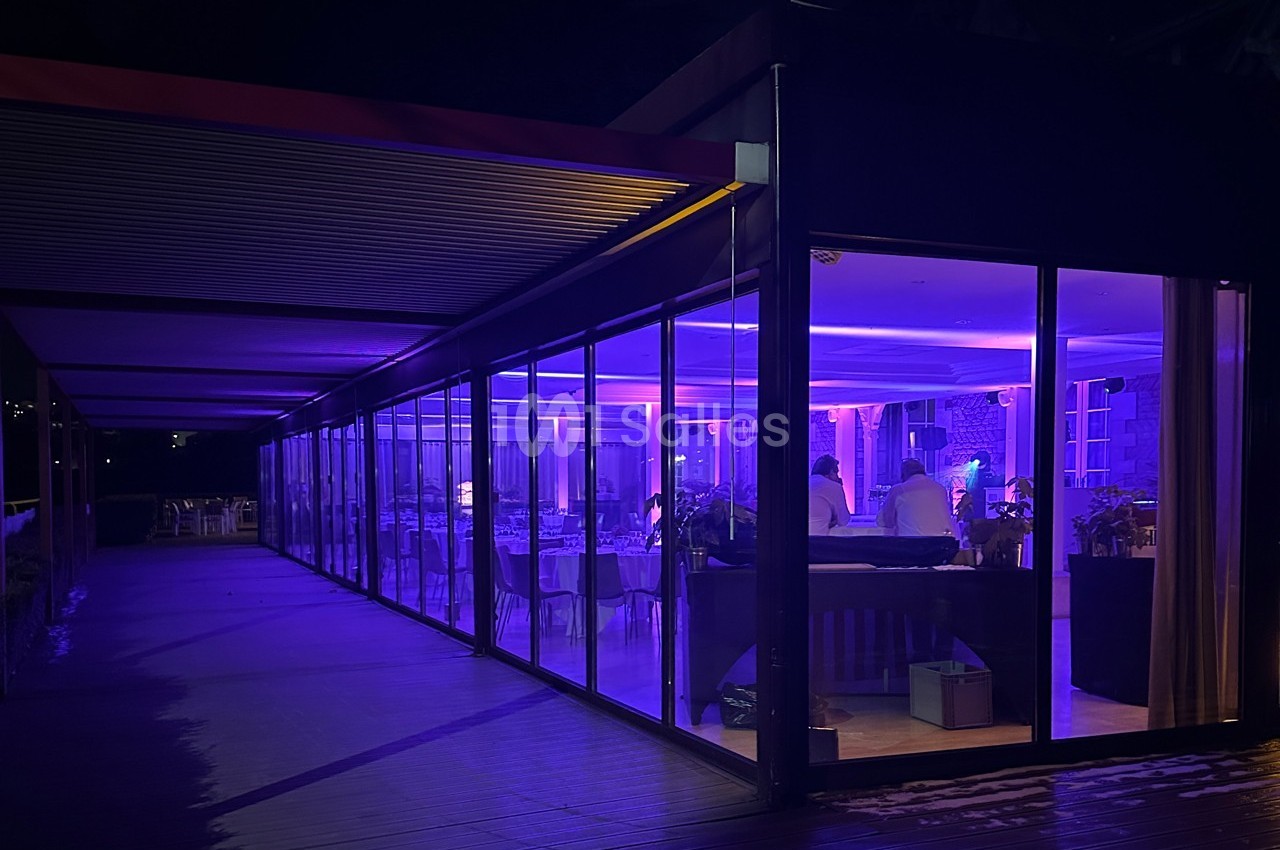 Salle vitrée éclairée en violet la nuit, avec des tables dressées et des personnes à l'intérieur.