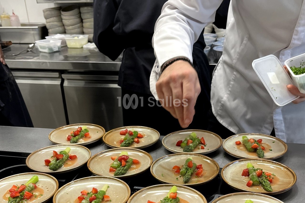 Un chef dispose des assiettes garnies d'asperges, fraises et herbes dans une cuisine professionnelle.