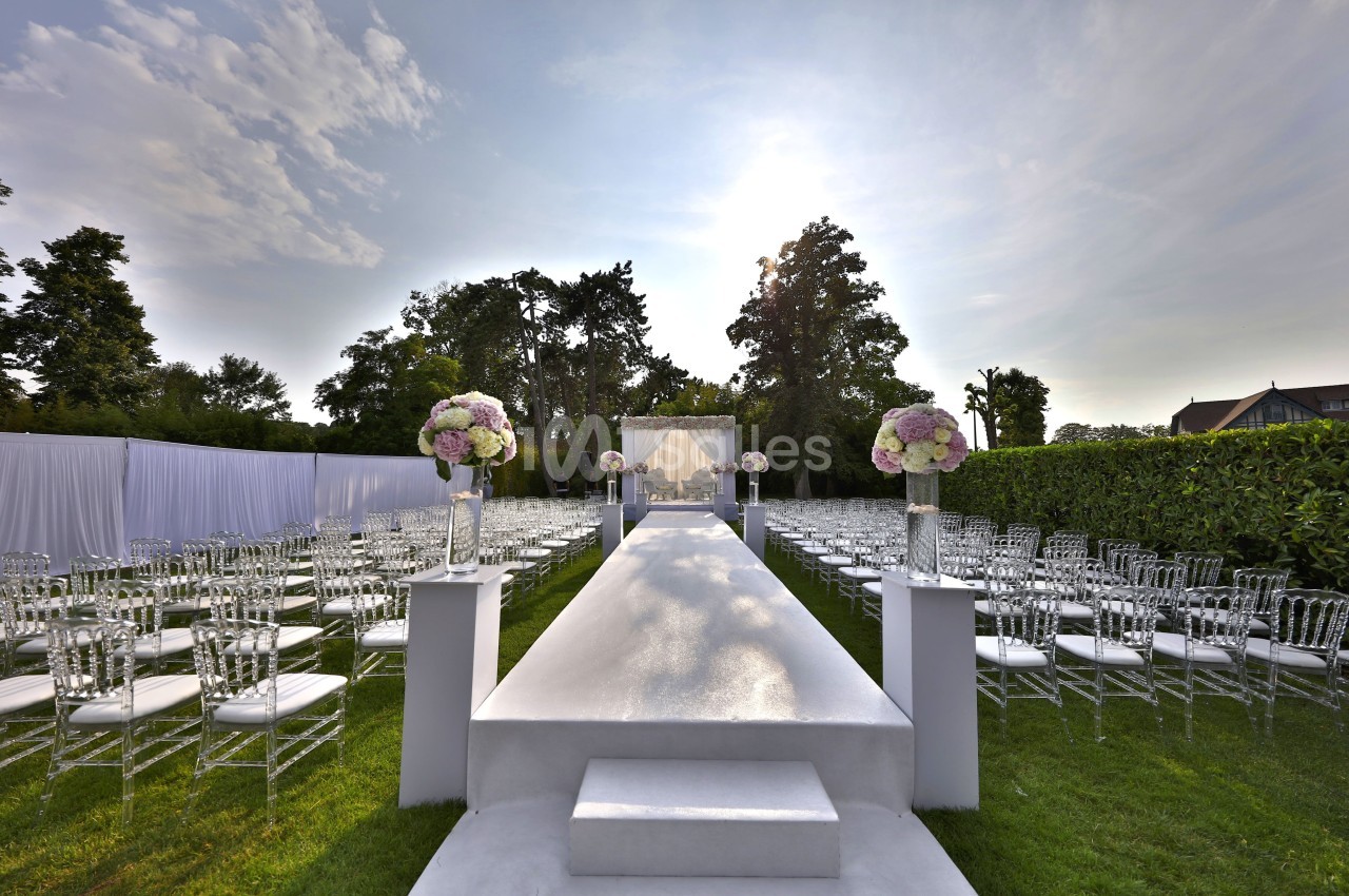 Allée centrale blanche bordée de chaises transparentes et de bouquets, menant à une arche de mariage en extérieur.