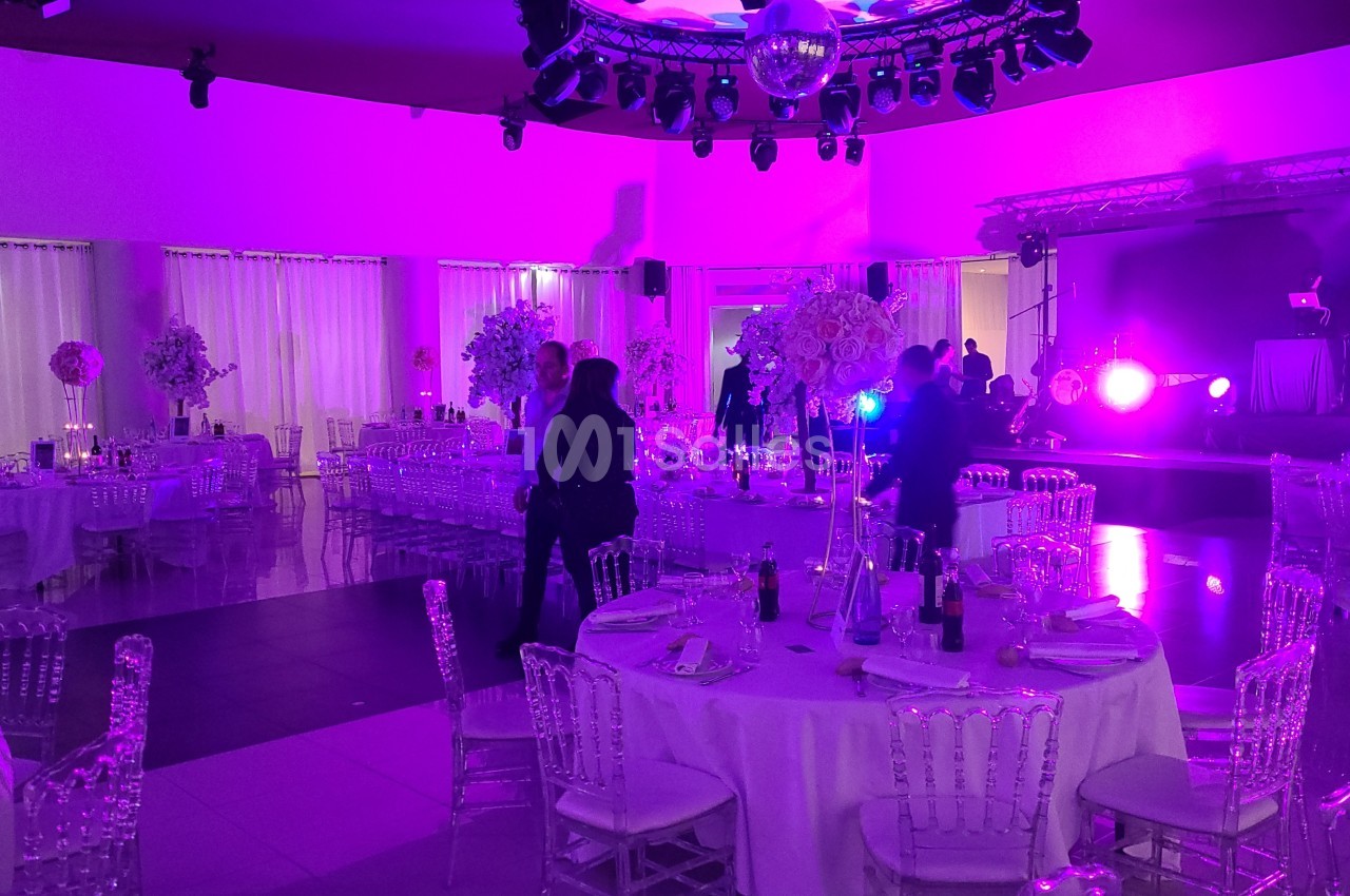 Salle de réception décorée avec des tables rondes, des chaises transparentes et un éclairage violet tamisé.