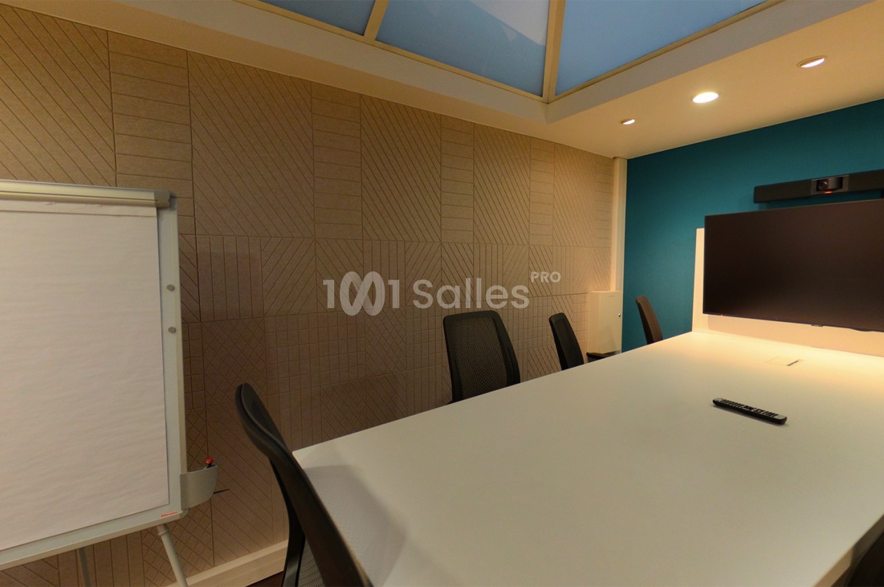 Salle de réunion moderne avec table blanche, chaises noires, écran mural, caméra de visioconférence et tableau blanc.
