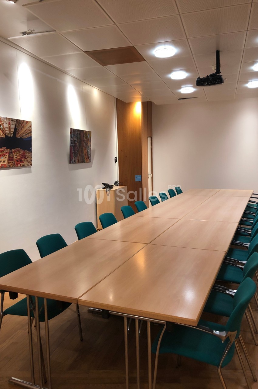 Salle de réunion avec une grande table rectangulaire en bois, entourée de chaises vertes, éclairée par des plafonniers.
