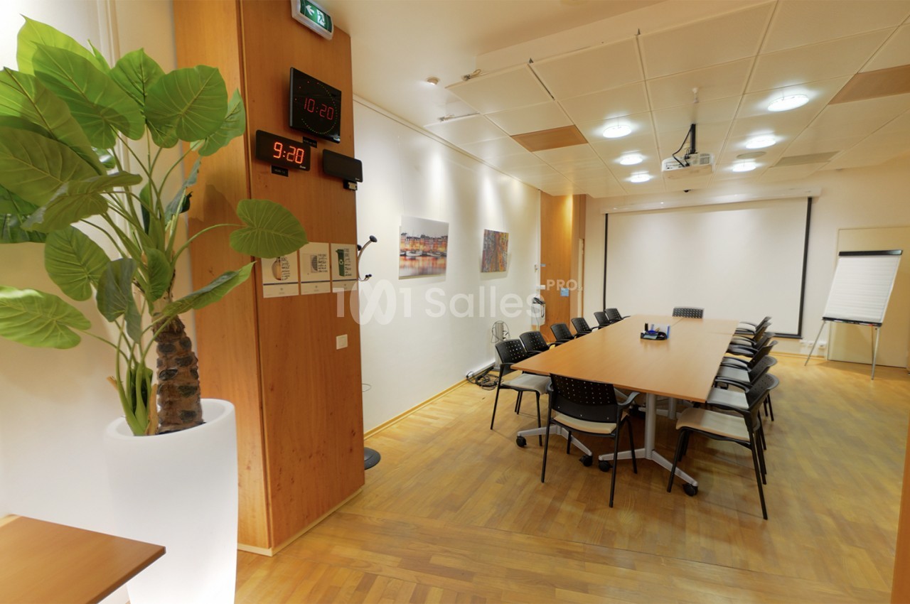Salle de réunion lumineuse avec table rectangulaire, chaises, tableau blanc, horloge murale et plante décorative.