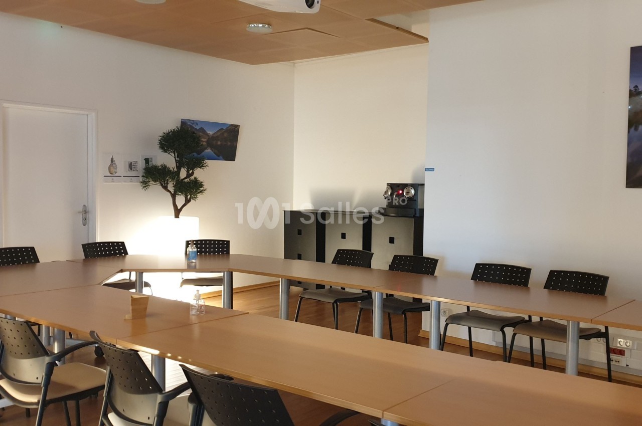 Salle de réunion lumineuse avec tables disposées en U, chaises noires, plantes et décorations murales.