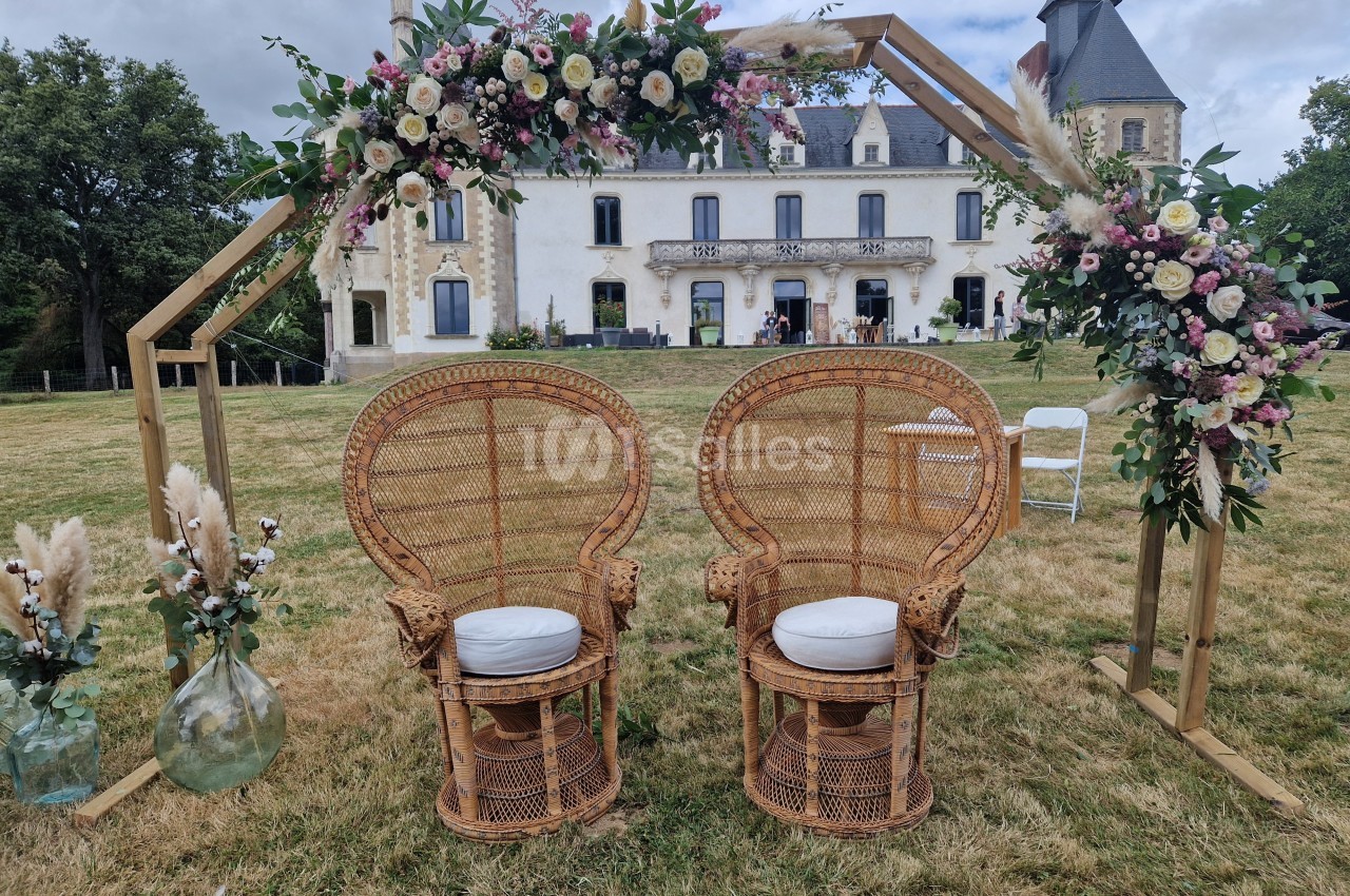 Deux fauteuils en rotin sous une arche fleurie, installés devant un château sur une pelouse.