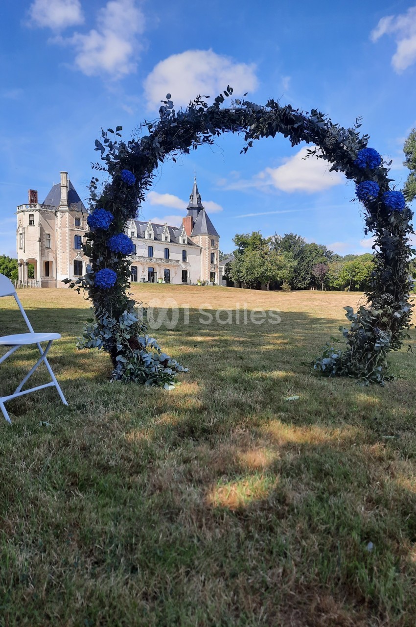 Arc fleuri avec des hortensias bleus sur une pelouse, devant un château sous un ciel bleu.