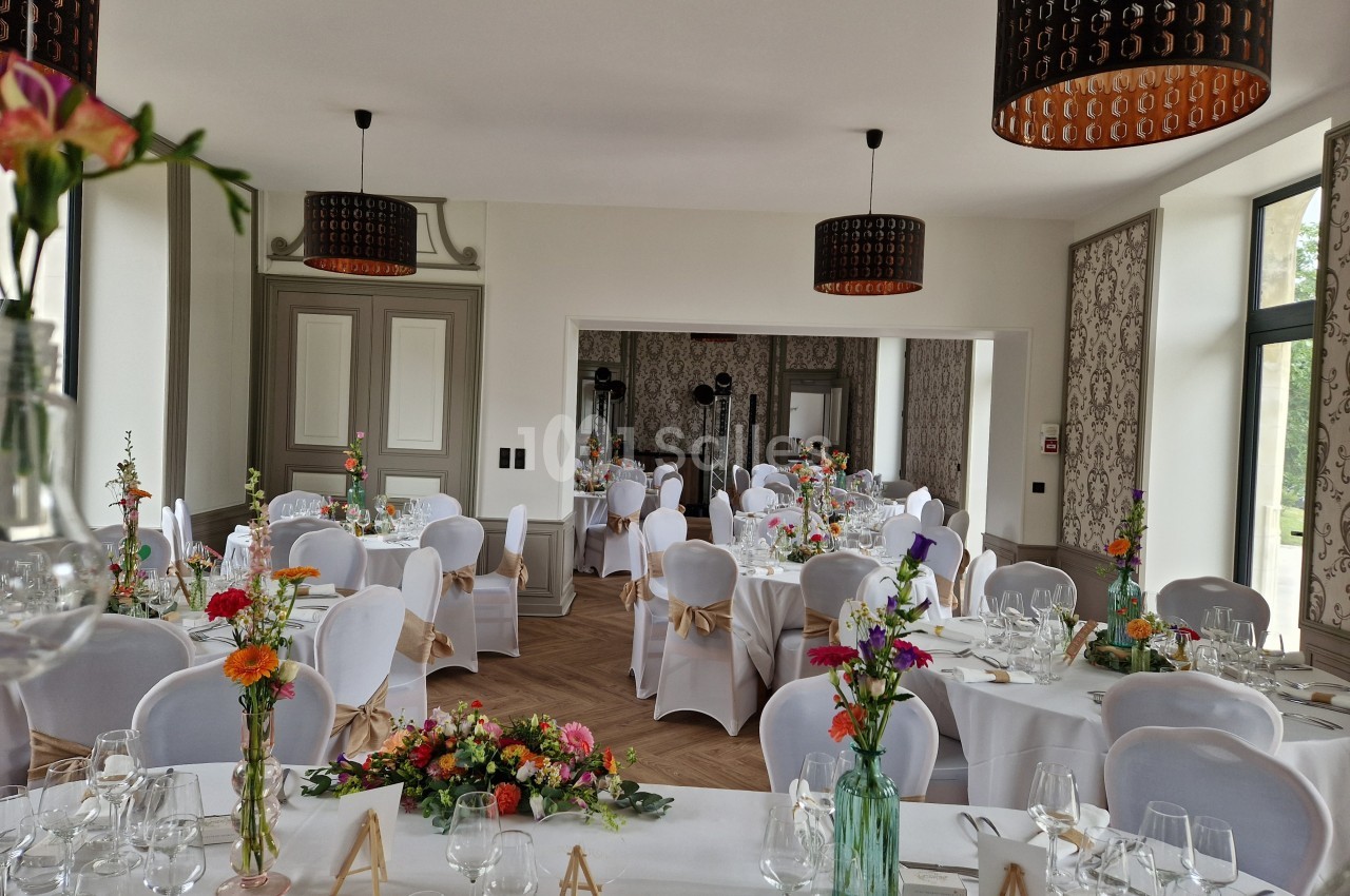 Salle de réception décorée avec des tables dressées, nappes blanches, fleurs colorées et lumière naturelle.