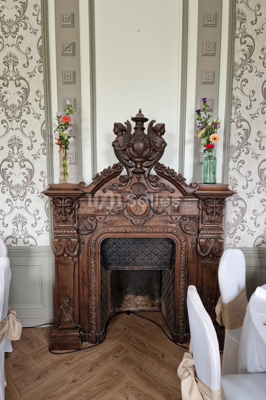 Cheminée en bois sculpté ornée de motifs décoratifs, entourée de chaises blanches et de vases fleuris.
