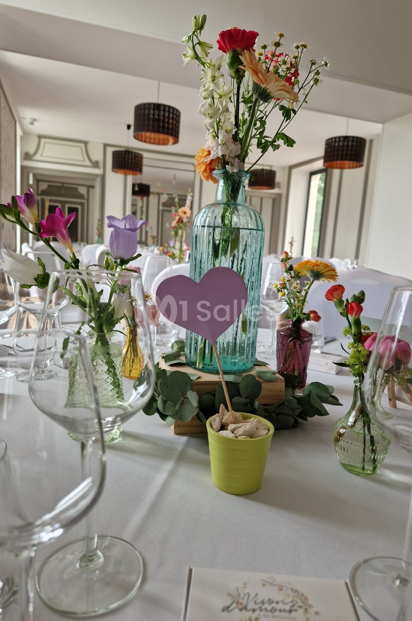 Table décorée pour un événement avec fleurs colorées, vases en verre, et un cœur violet planté dans un pot vert.
