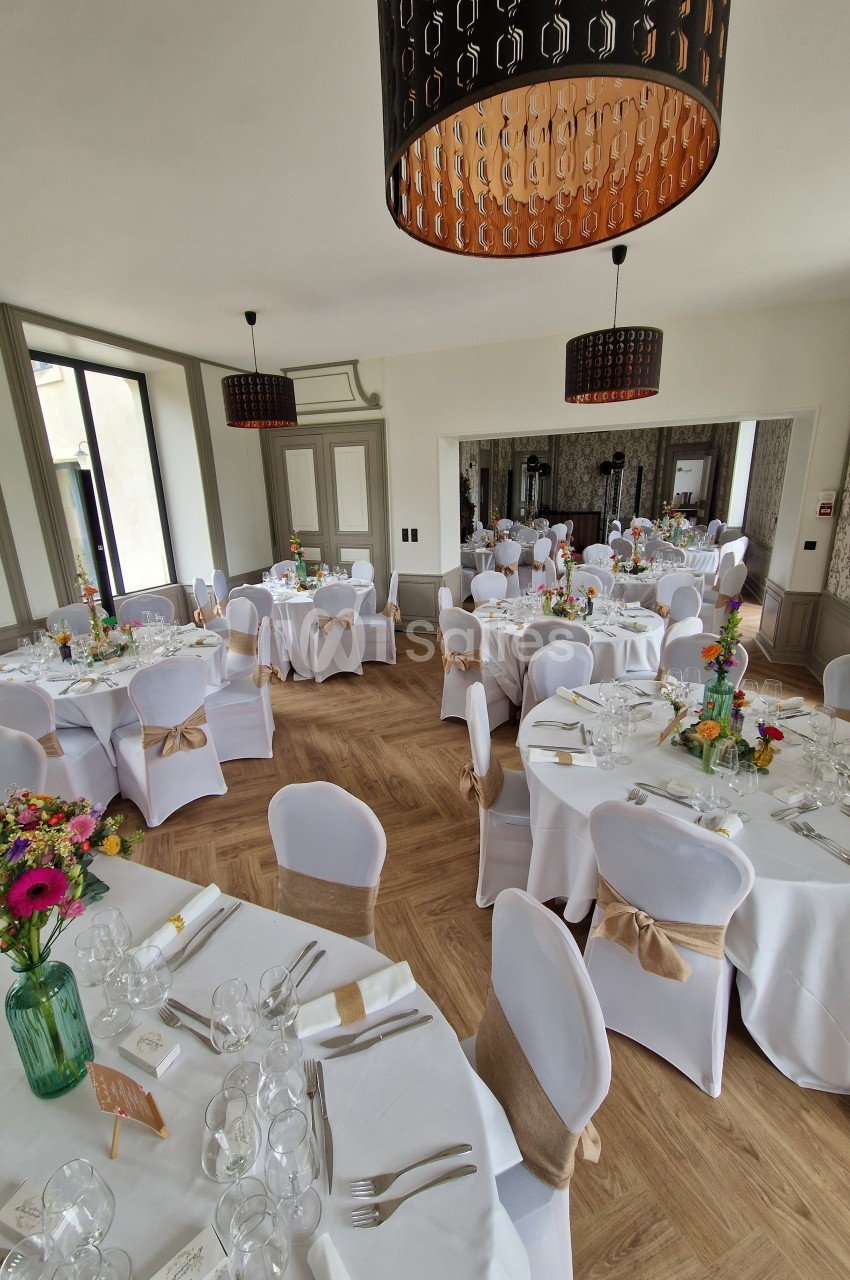 Salle de réception lumineuse avec tables rondes décorées de nappes blanches, fleurs colorées et chaises ornées de nœuds…