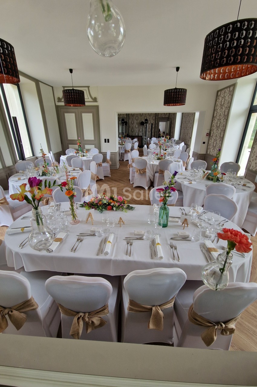 Salle de réception décorée pour un événement, avec tables dressées, nappes blanches et bouquets de fleurs colorées.