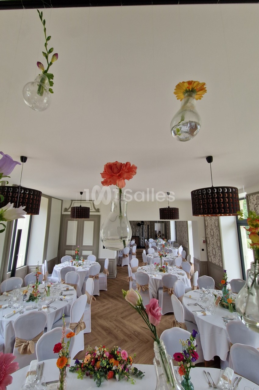 Salle de réception décorée avec des tables dressées et des fleurs suspendues dans des vases en verre.