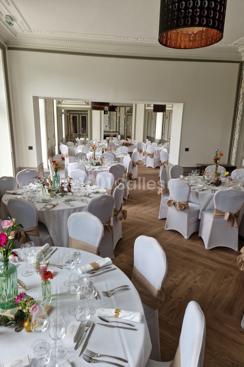 Salle de réception élégante avec tables rondes dressées, nappes blanches et décorations florales colorées.