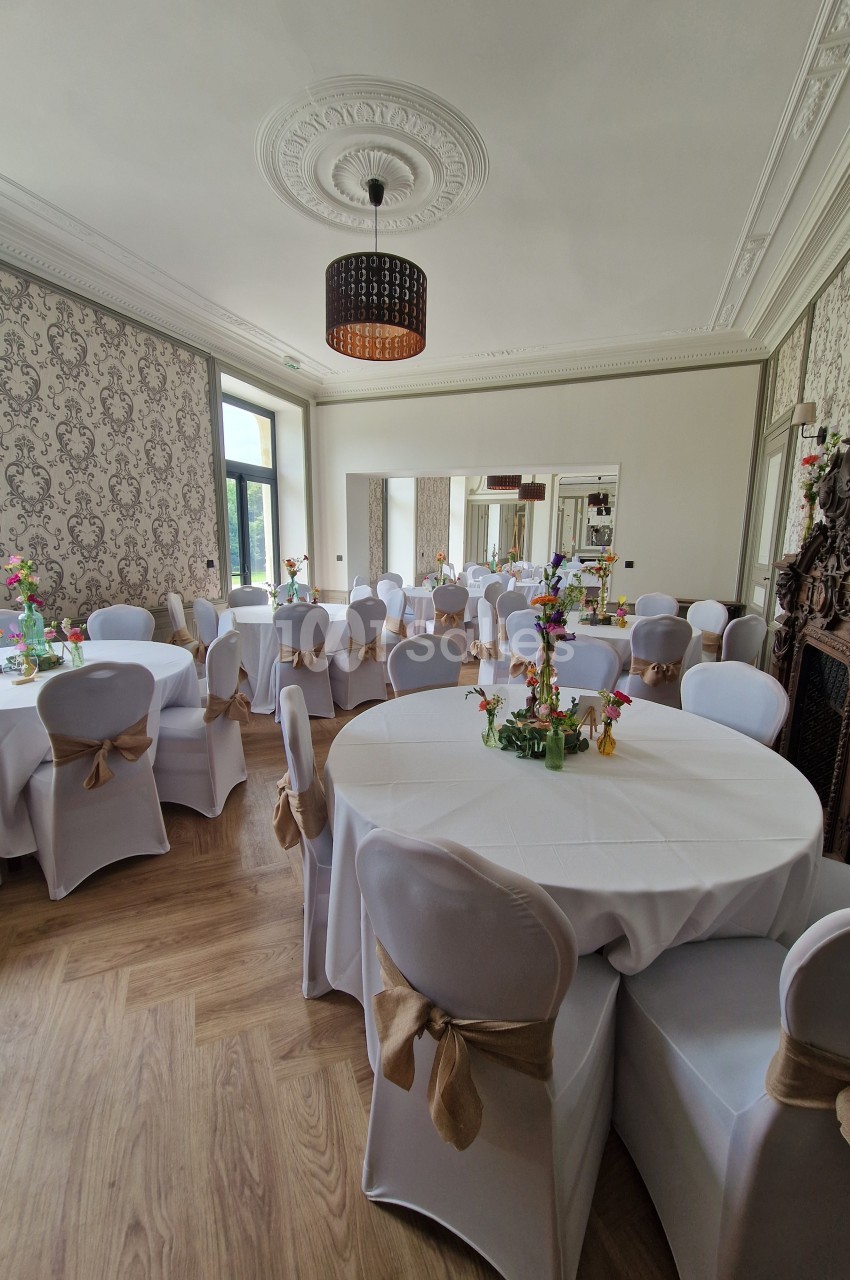 Salle élégante avec tables rondes dressées, nappes blanches, chaises décorées et lumière naturelle entrant par les fenêtres.