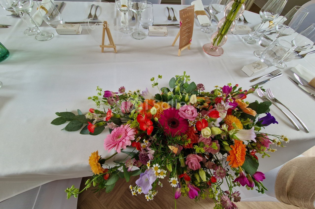 Centre de table fleuri aux couleurs vives sur une nappe blanche, entouré de couverts et de verres élégamment disposés.