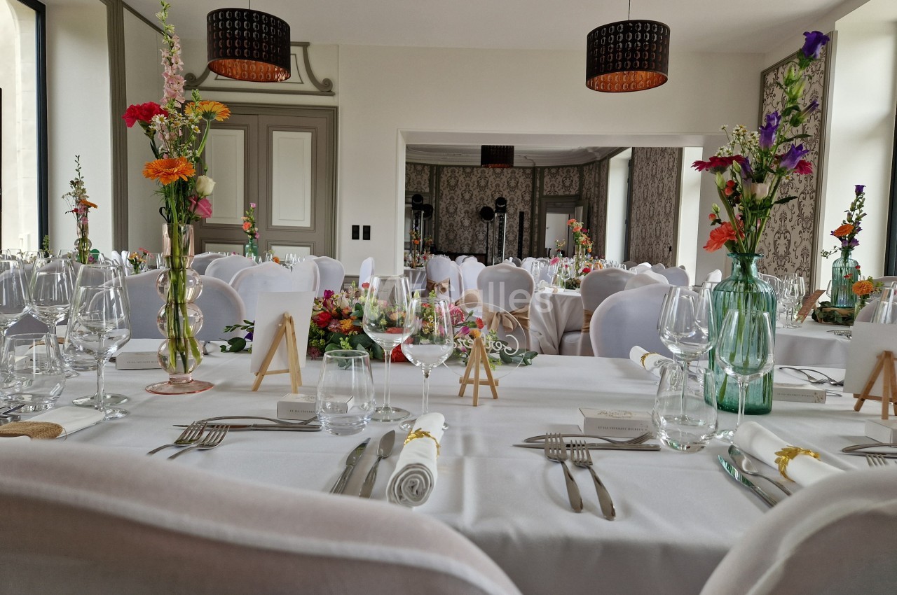 Salle de réception décorée avec des tables dressées, nappes blanches, fleurs colorées et chaises blanches.