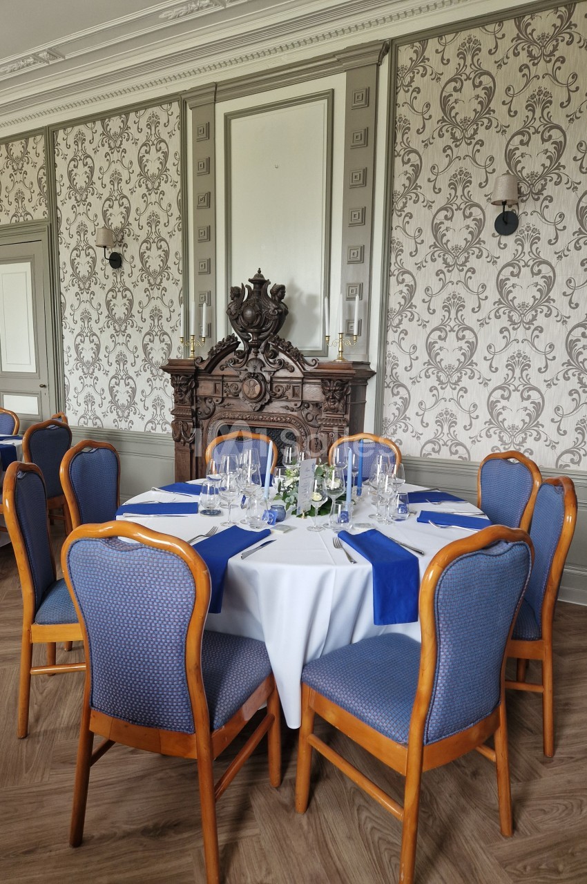 Salle élégante avec table ronde dressée, nappes blanches et bleues, chaises rembourrées, et cheminée décorative en arrière…