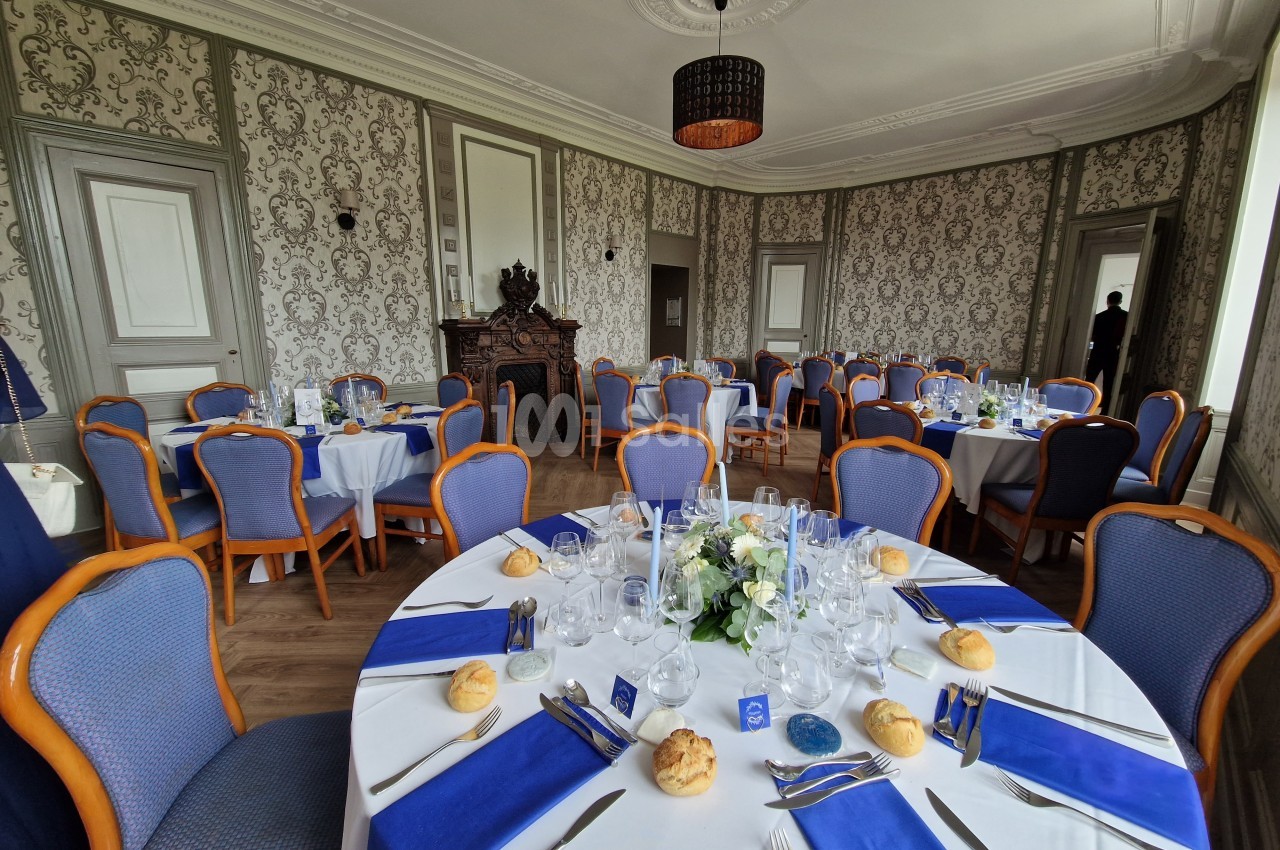 Salle de réception élégante avec tables dressées, nappes blanches, serviettes bleues et décoration florale centrale.
