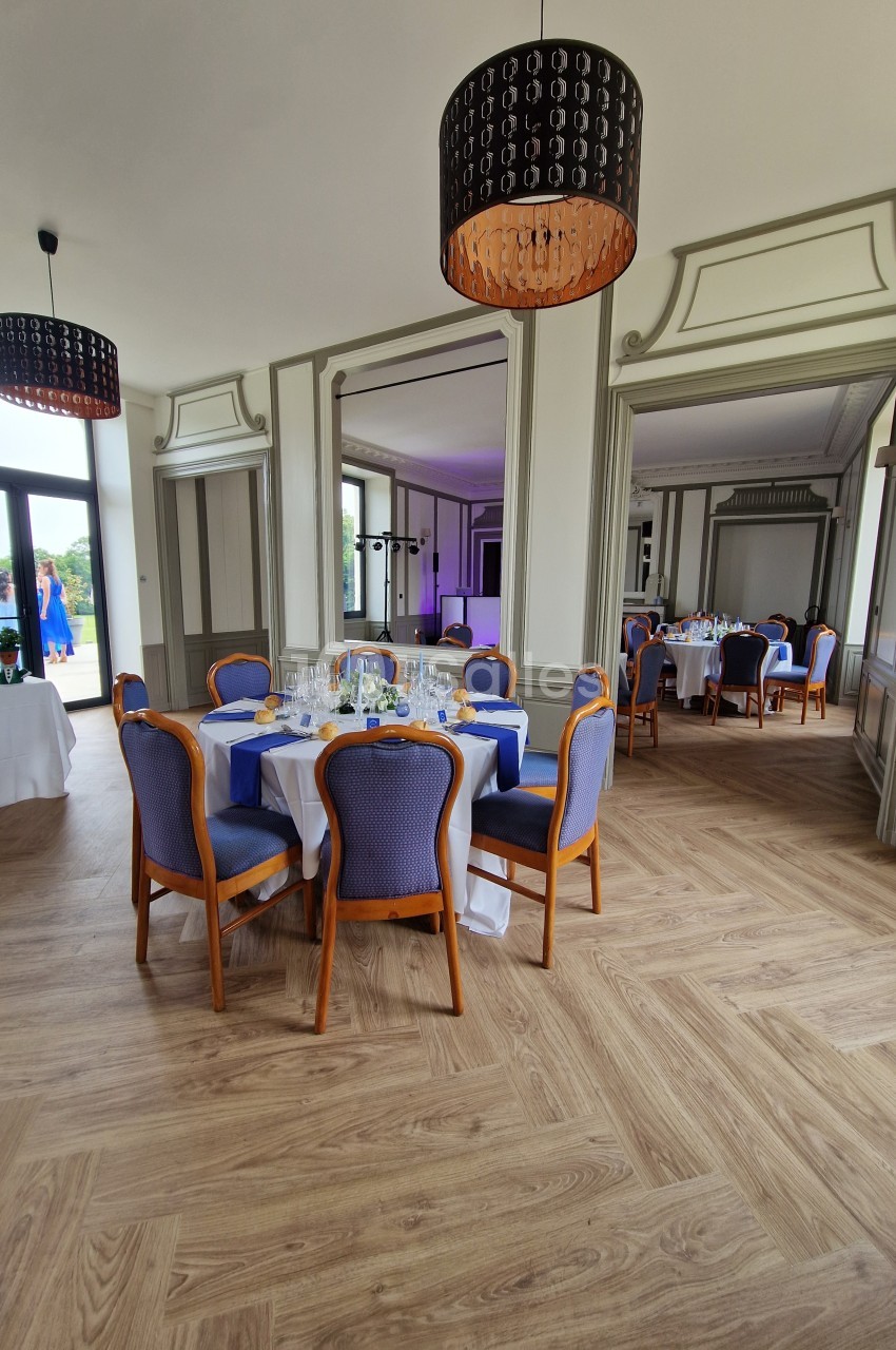 Salle élégante avec tables dressées, chaises bleues et luminaires suspendus, parquet clair et grandes fenêtres.